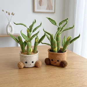 May include: Two crocheted bamboo plants in round pots. One pot is cream with tan feet, the other is tan with dark brown feet. Both pots feature embroidered faces with black eyes. The bamboo stalks are green with textured segments and pointed leaves.