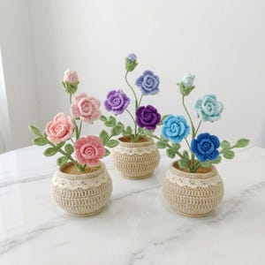 Puede incluir: Tres arreglos florales de ganchillo en macetas de hilo beige con borde de encaje blanco. Cada maceta contiene tallos con hojas verdes y rosas en tonos rosa, morado y azul, incluyendo varios capullos pequeños en tallos verdes finos.