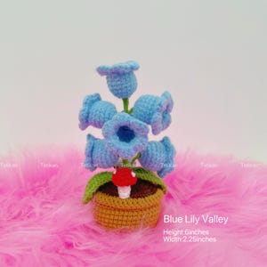 May include: A crocheted Blue Lily Valley arrangement. The arrangement features six light blue bell-shaped flowers, green leaves, and a red and white mushroom, all in a woven basket. The height is 6 inches, and the width is 2.25 inches.