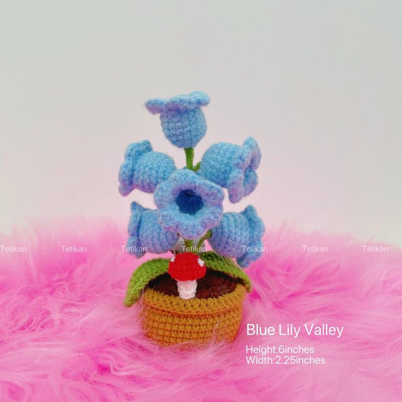 May include: A crocheted Blue Lily Valley arrangement. The arrangement features six light blue bell-shaped flowers, green leaves, and a red and white mushroom, all in a woven basket. The height is 6 inches, and the width is 2.25 inches.
