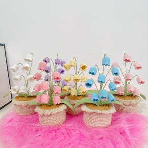 Może przedstawiać: Kolekcja ręcznie robionych szydełkowych konwalii w małych doniczkach. Każda doniczka ma inny kolor kwiatów, w tym biały, różowy, fioletowy, żółty i niebieski. Doniczki są jasnoróżowe z falistą krawędzią.