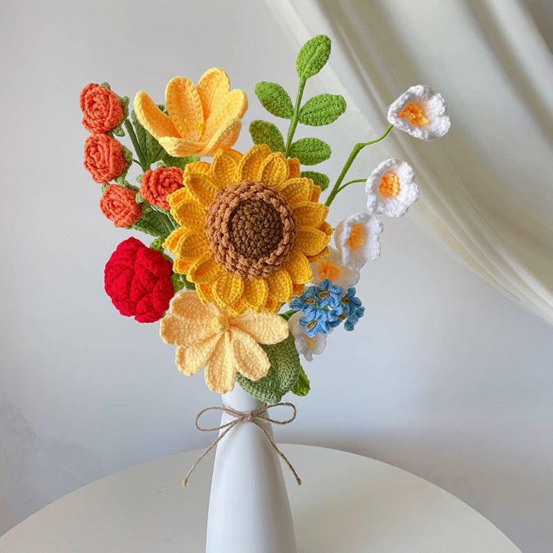 This is a sunflower-themed crochet bouquet placed in a white vase. There are a total of 8 stems of flowers in the bouquet, including 1 stem of double-layer yellow petals sunflower, 1 stem of yellow tulip, 1 stem of white multi-headed lily of the valley, 1 stem of orange multi-headed rose, 1 stem of red rose, 1 stem of yellow geranium, 1 stem of blue forget-me-not, and 1 stem of eucalyptus leaf. The leaves and stems of the crochet flowers are green. The bouquet is placed on the table.