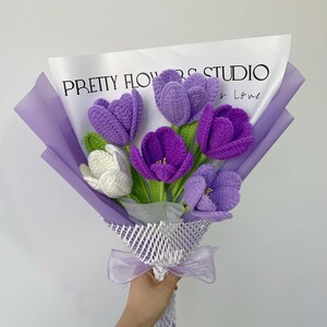 Pu&ograve; includere: Un mazzo di fiori all'uncinetto viola e bianchi avvolti in carta velina viola e rete bianca con un nastro viola. Il mazzo &egrave; etichettato "PRETTY FLOWERS STUDIO & LOVE".