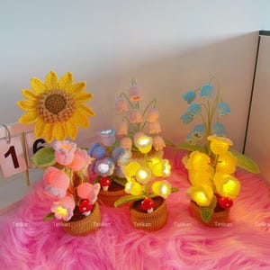 Peut inclure: Plusieurs lampes en crochet en forme de fleurs, de couleurs jaune, rose et bleu. Chaque lampe est posée sur un socle en forme de panier tressé et ornée d'un petit champignon. Les lampes sont éclairées.