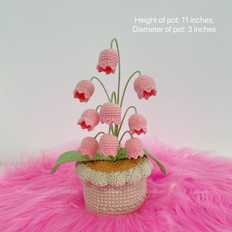 May include: A handmade crochet flower arrangement featuring pink bell-shaped flowers with green stems and leaves. The flowers are in a beige pot with a scalloped edge. The pot is 11 inches tall and 3 inches in diameter. The text "Tetikan" is visible.