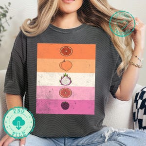 Pode incluir: T-shirt cinzenta escura com um design colorido de frutas. O design inclui fatias de toranja, um pêssego, fruta do dragão e amora, dispostas em faixas horizontais. A t-shirt é de estilo casual.