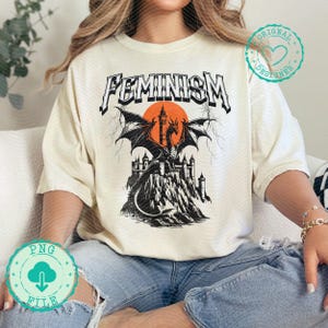 Pode incluir: Uma camiseta branca com um gráfico preto e branco de um dragão na frente de um castelo. O texto "FEMINISM" é impresso em preto acima do gráfico.