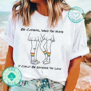 Pode incluir: Camiseta branca com o texto "BE CAREFUL WHO YOU HATE IT COULD BE SOMEONE YOU LOVE". A camiseta apresenta um desenho de duas figuras com meias arco-íris. Um logotipo de arquivo PNG é visível.