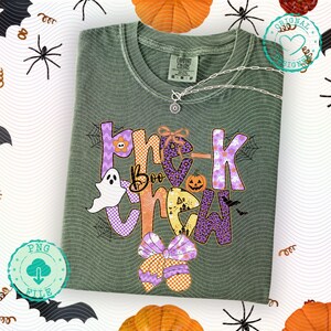 May include: A sage green t-shirt with a colorful Halloween-themed graphic that says "Trick or Treat Boo Crew". The design includes a ghost, pumpkins, bows, and spiderwebs. A silver necklace with a pendant is draped on the shirt.