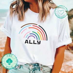 Puede incluir: Camiseta blanca con un diseño de arcoíris colorido y la palabra "ALLY" en letras negras. El arcoíris presenta varios colores, incluyendo marrón, rosa, naranja, verde y azul. La camiseta tiene las mangas dobladas.