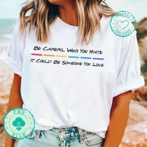 Pode incluir: Camiseta branca com mangas enroladas. A camiseta apresenta o texto "BE CAREFUL WHO YOU HATE IT COULD BE SOMEONE YOU LOVE" em preto, com uma linha de arco-íris abaixo da primeira linha de texto. Um logotipo de arquivo PNG está no canto inferior esquerdo.