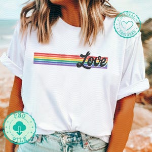 May include: White t-shirt featuring a rainbow-colored horizontal stripe design with the word "Love" in a black script font. The shirt has rolled-up sleeves and is made of a soft material.