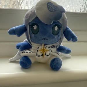 May include: A blue and white plush toy with a star on its chest. The toy has a white hood with a blue and white pattern. The toy is sitting on a white surface.