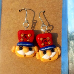 May include: A pair of dangle earrings featuring a whimsical clay figurine of a man with a red hat, blue collar, and a moustache. The figurine is painted with details like eyes, a smile, and a red and gold hat.