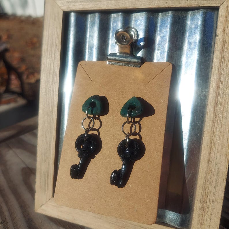 May include: A pair of dangle earrings with a dark green triangle charm and a black key charm hanging from a silver chain.