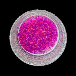 May include: A clear glass jar filled with vibrant pink and magenta glitter. The glitter is densely packed, creating a textured appearance. The jar has a decorative, etched design around the rim, set against a black background.