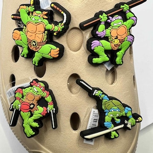 Può includere: Quattro ciondoli colorati per scarpe delle Tartarughe Ninja. Ogni ciondolo presenta un diverso personaggio di tartaruga in pose d'azione, con accenti verdi, arancioni, rossi e blu. I ciondoli sono attaccati a una scarpa beige.
