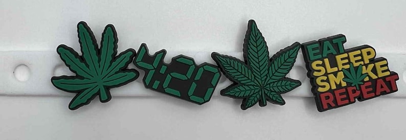 May include: Four shoe charms with cannabis leaf designs and the text "4:20" and "EAT SLEEP SMOKE REPEAT". The charms are green and black, with a white background.