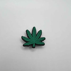 May include: A green cannabis leaf-shaped charm with a black outline. The charm is a vibrant green color and has detailed leaf veins. The charm is likely made of rubber or a similar material. The background is plain white.