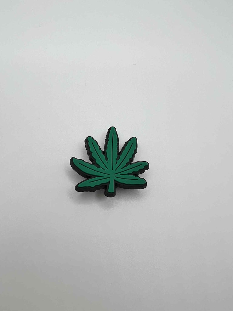 May include: A green cannabis leaf-shaped charm with a black outline. The charm is a vibrant green color and has detailed leaf veins. The charm is likely made of rubber or a similar material. The background is plain white.