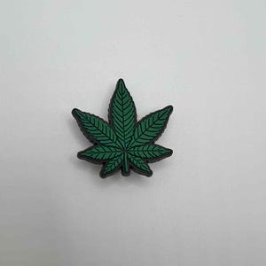 May include: A green cannabis leaf charm with black trim. The leaf features detailed veining and is set against a white background. This decorative charm is suitable for various craft projects.