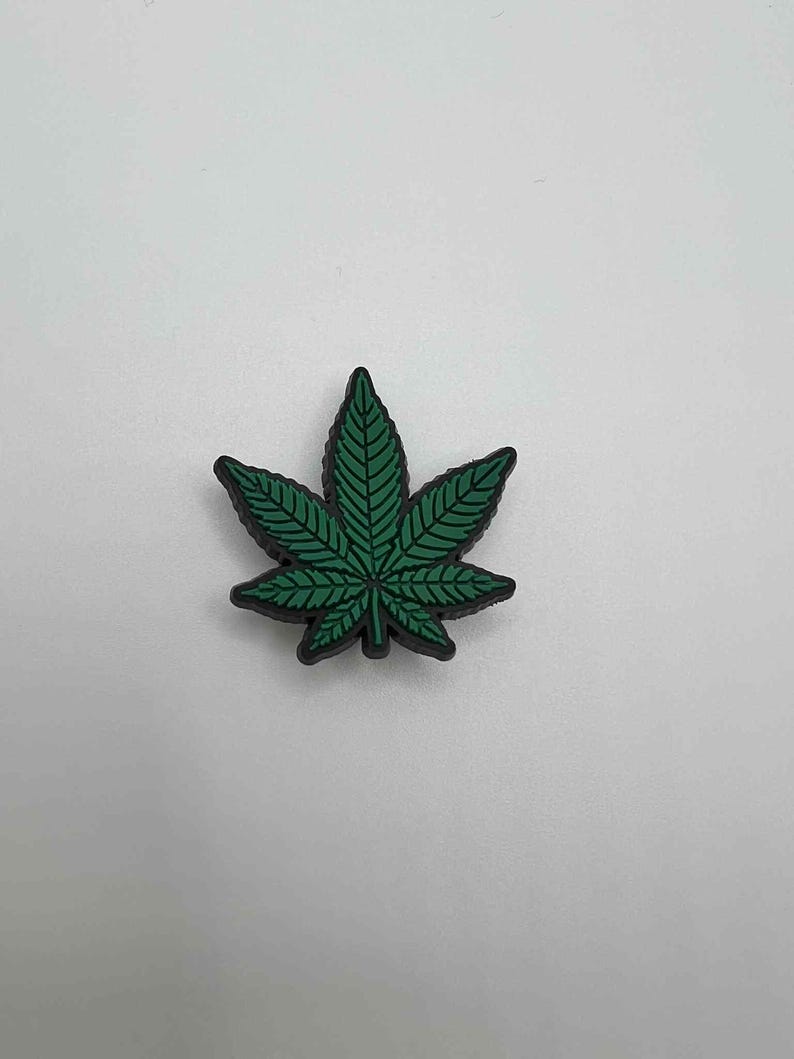 May include: A green cannabis leaf charm with black trim. The leaf features detailed veining and is set against a white background. This decorative charm is suitable for various craft projects.