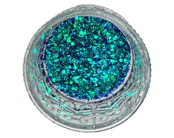 Puede incluir: Un tarro de vidrio transparente lleno de purpurina azul turquesa y azul vibrante. La purpurina es una mezcla de partículas pequeñas y medianas, creando un efecto brillante. El tarro tiene un diseño decorativo grabado alrededor del borde.
