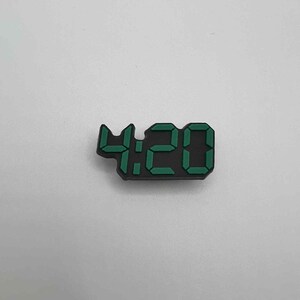 May include: A black digital clock charm displaying the time 4:20 in green. The charm is a small, rectangular shape, with the numbers clearly visible against the black background. The numbers are in a digital font.