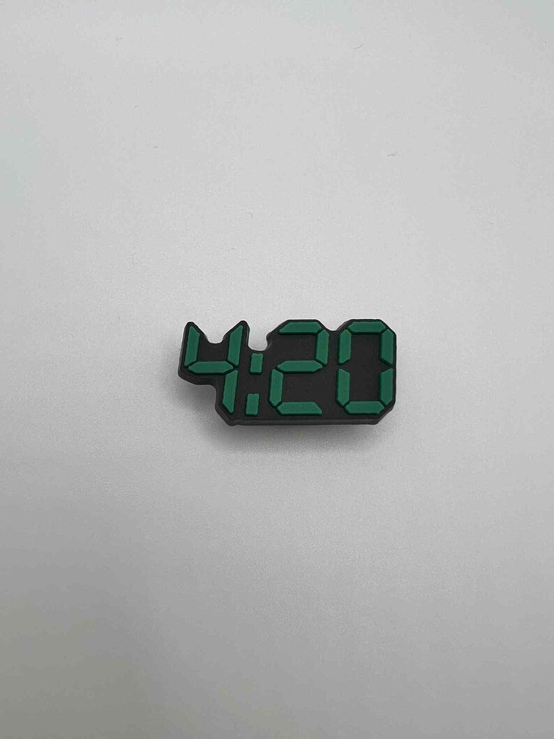 May include: A black digital clock charm displaying the time 4:20 in green. The charm is a small, rectangular shape, with the numbers clearly visible against the black background. The numbers are in a digital font.