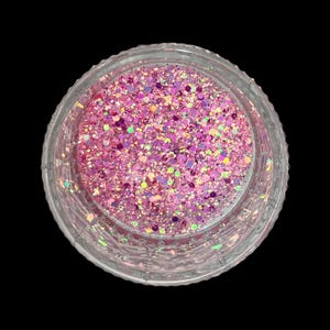 May include: A clear, round container filled with pink glitter. The glitter is a mix of various shapes and sizes, with iridescent and holographic elements. The container is set against a black background.