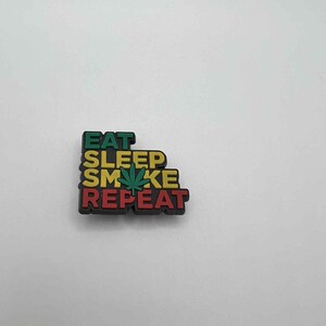 May include: A colorful shoe charm with the words "EAT SLEEP SMOKE REPEAT" in green, yellow, and red. A green cannabis leaf is in the center of the word "SMOKE". The charm is on a white background.