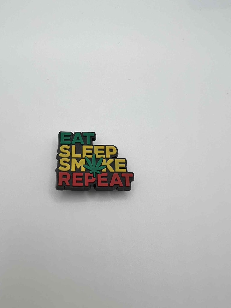 May include: A colorful shoe charm with the words "EAT SLEEP SMOKE REPEAT" in green, yellow, and red. A green cannabis leaf is in the center of the word "SMOKE". The charm is on a white background.