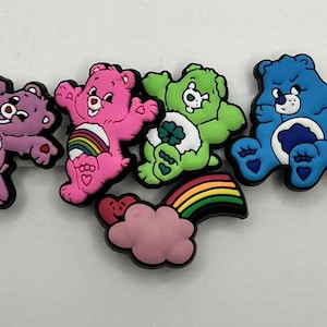 Op de afbeelding: Een verzameling kleurrijke Care Bear schoenbedels. De bedels tonen verschillende beren in paars, roze, groen, blauw en geel, elk met unieke buikbadges. Een regenboog- en wolkbedel is ook inbegrepen.
