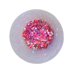 Puede incluir: Un cuenco transparente y decorativo lleno de un grupo de pequeños brillantes redondos rosas y plateados. El cuenco tiene un borde festoneado y está sobre un fondo blanco. Los brillantes están densamente empaquetados, creando un efecto brillante.