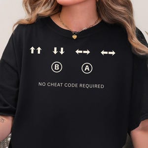 May include: Black t-shirt with white graphic of up, down, left, and right arrows, and A and B buttons. The text "NO CHEAT CODE REQUIRED" is below the graphic. The shirt is a casual, oversized fit.