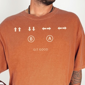 Op de afbeelding: Een roestkleurig t-shirt met witte richtingtekens en de tekst "GIT GOOD". Het shirt heeft een vintage uitstraling. Een voetbal wordt in de arm van de persoon gehouden. Het t-shirt is een casual kledingstuk.