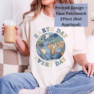 May include: Cream-colored t-shirt with a round graphic of the Earth and the words "Earth Day Every Day" in a blue and green font. The shirt has a relaxed fit and is paired with blue jeans.