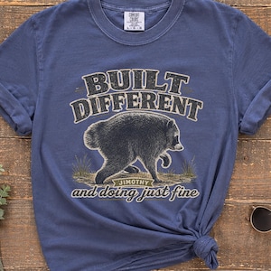 May include: A blue short-sleeve t-shirt featuring a graphic of a black bear walking on its hind legs. The text above the bear reads "BUILT DIFFERENT" and the text below reads "JIMOTHY" and "and doing just fine" in a stylized font.