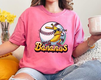 May include: Pink t-shirt with a cartoon banana wearing sunglasses and holding a baseball bat. The banana is standing on a baseball. The text "Bananas" is written in a yellow, curved font below the banana.