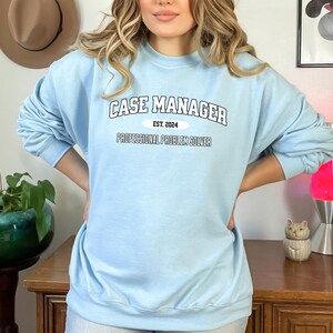 May include: Light blue sweatshirt with the text "CASE MANAGER EST. 2024 PROFESSIONAL PROBLEM SOLVER" printed on the front.
