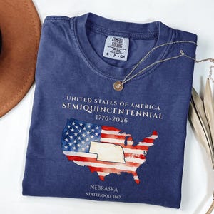 Op de afbeelding: Een marineblauw t-shirt met een kaart van de Verenigde Staten in het ontwerp van de Amerikaanse vlag. De tekst op het shirt luidt "UNITED STATES OF AMERICA SEMIQUINCENTENNIAL 1776-2026" en "NEBRASKA STATEHOOD: 1867". Een gouden ketting ligt over het shirt.