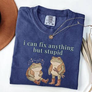 May include: A navy blue t-shirt featuring the phrase "i can fix anything but stupid" and a graphic of two frogs in cowboy boots and vests. A gold necklace is draped across the shirt.