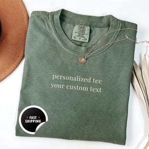 Könnte beinhalten: Ein olivgrünes T-Shirt mit dem Text "personalized tee your custom text" auf der Vorderseite. Eine goldene Halskette mit Anhänger liegt auf dem Shirt. Ein "Fast Shipping"-Aufkleber befindet sich unten links.