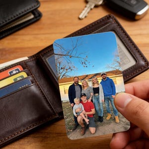 May include: A rectangular metal wallet card featuring a family photo, held near a brown leather wallet with visible credit cards and cash. The background includes keys and a car remote on a wooden surface. The card displays a family portrait under a blue sky.