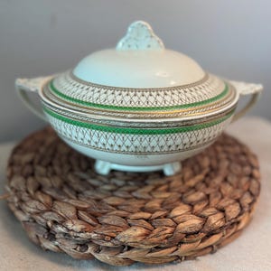 May include: A white ceramic serving bowl with a lid, featuring a green and gold geometric pattern around the rim. The bowl has two handles and sits on a woven placemat. The lid has a decorative finial.