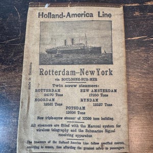 May include: A rectangular beige fabric document featuring a black printed illustration of a steamship. The text reads Holland-America Line, Rotterdam-New York via Boulogne-Sur-Mer, listing twin screw steamers and their tonnage, plus technical details.