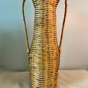 May include: A tall, woven wicker vase with two curved handles. The vase is cylindrical with a flared top and a rounded base. The natural tan color of the wicker is visible throughout the piece, showcasing the intricate weaving pattern. A decorative home accent.