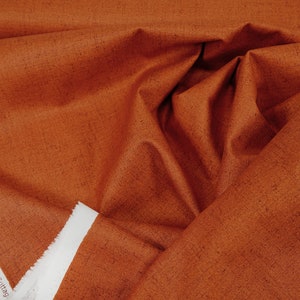 Puede incluir: Primer plano de una tela de color óxido con una textura sutilmente moteada. La tela está doblada y muestra el borde de la orilla con el texto "Copyright of Needl' Times for Andover Fabrics © 2022 Patti Cazeault Cottage".