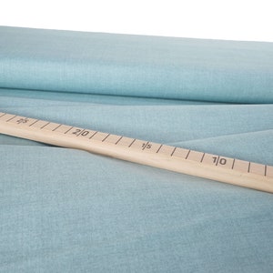 Peut inclure: Un tissu bleu clair drapé, avec une règle en bois posée dessus. La règle est marquée de lignes et de chiffres noirs, indiquant les mesures en centimètres. Le tissu a un aspect texturé.