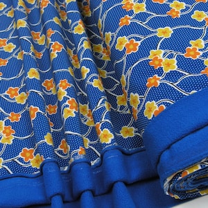 Op de afbeelding: Blauwe stof met een wit en oranje bloemenpatroon. De stof is gevouwen en opgerold, waardoor de blauwe onderkant zichtbaar is.